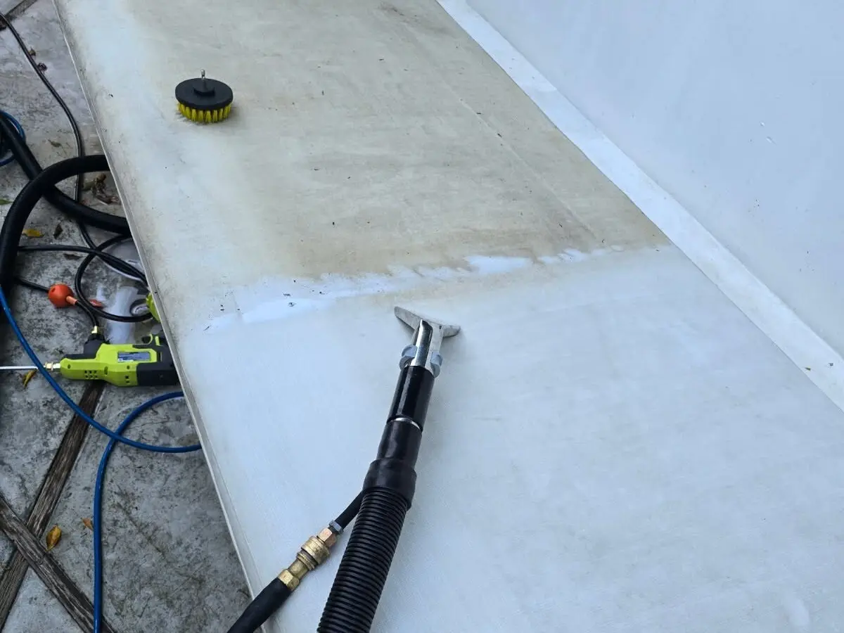 Photo of a long white outdoor sofa cushion mid-cleaning: the upper half still brown with grime, the lower half clean, with an upholstery extraction tool and its hose on the cushion and a drill brush resting on the dirty half.