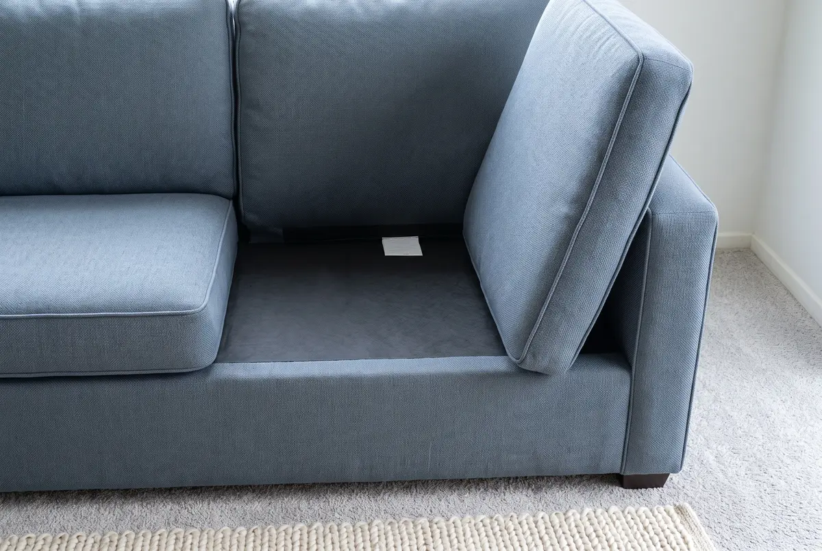Illustration of a slate-blue fabric sofa with one seat cushion lifted to show the small sewn-in care tag on the seat deck.